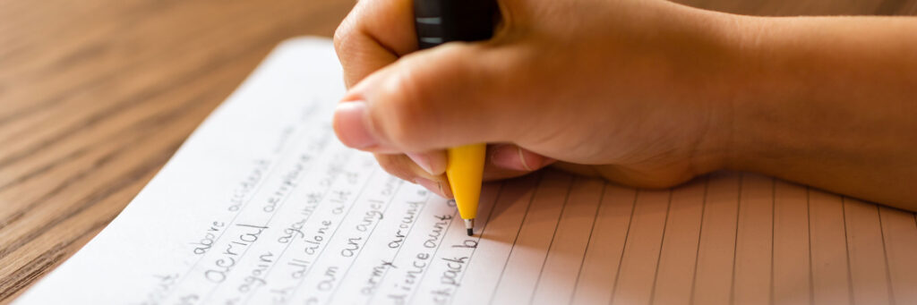 child's hand writing an essay in notebook with pencil